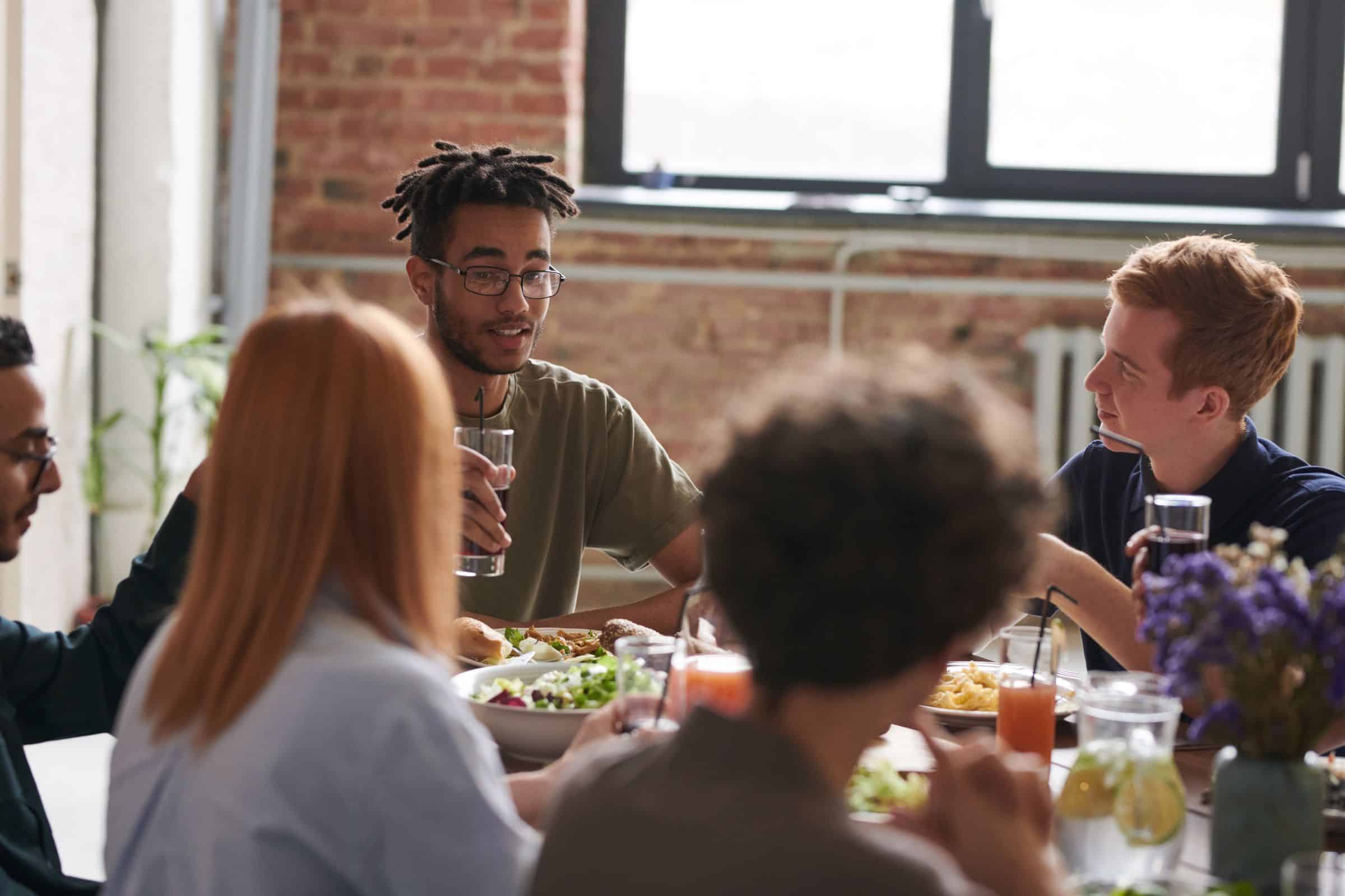 Young adults talking and eating together around a long wooden table in a sunny room