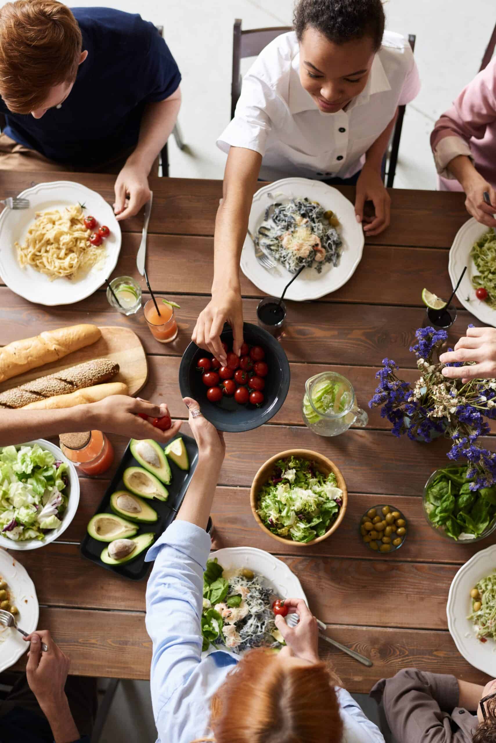 Overhead view of friends passing dishes of salad, pasta and bread around a shared wooden table